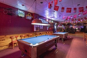 A pool table with a neon sign, a bar area, and a ceiling with hanging banners.