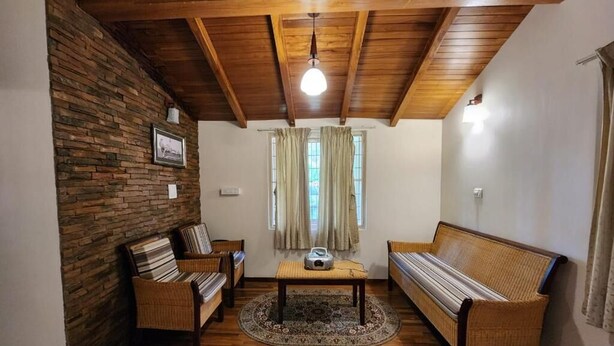 Una habitación con techo de madera, pared de piedra, sofá, una silla, una mesita y una ventana con cortinas.