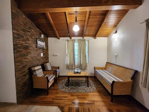Una habitación con techo de madera, pared de piedra, sofá, una silla, una mesita y una ventana con cortinas.