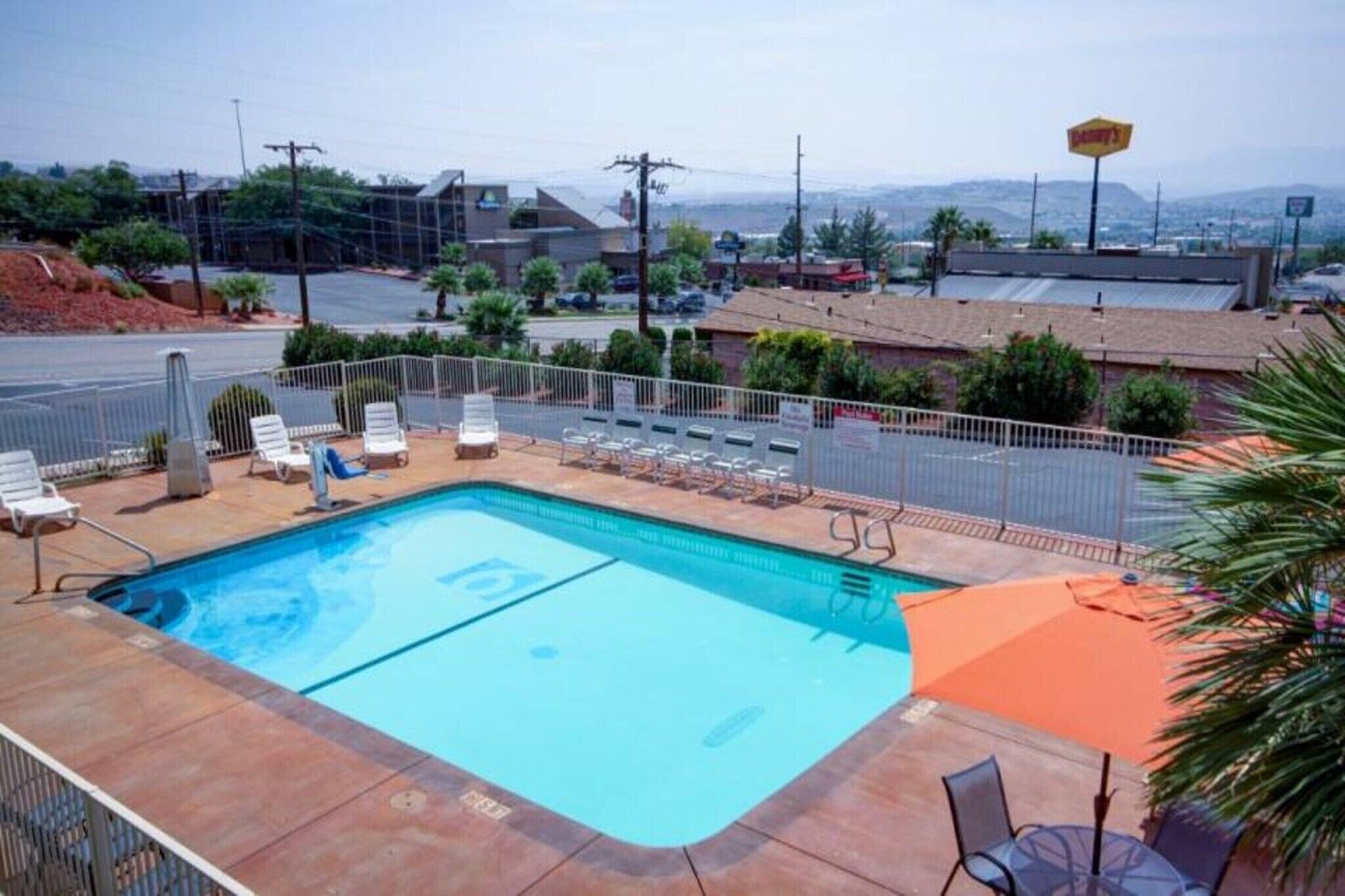 A swimming pool with lounge chairs and a fenced area, surrounded by buildings and a clear sky.