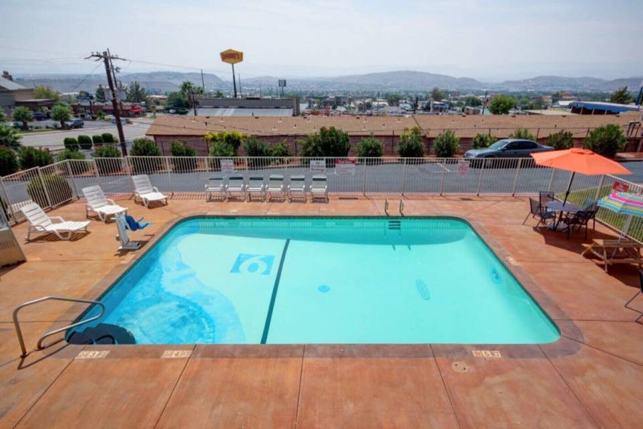 A rectangular swimming pool with a metal ladder, surrounded by a white fence. There are lounge chairs and a table with an umbrella on the poolside.