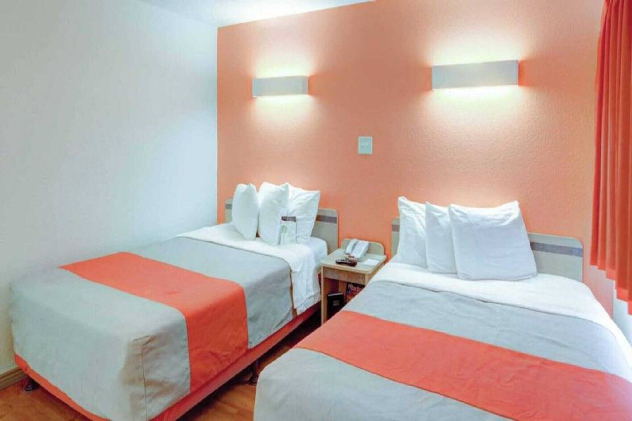 A hotel room with two beds, each with white bedding and orange accents, a small bedside table with a phone, and wall-mounted light fixtures.