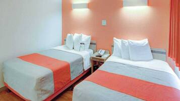 A hotel room with two beds, each with white bedding and orange accents, a small bedside table with a phone, and wall-mounted light fixtures.