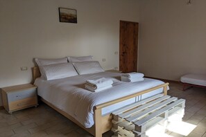 Ein ordentlich bezogenes Bett mit Kissen und gefalteten Handtüchern, eine hölzerne Bank, ein Nachttisch mit Schubladen und eine Holztür.