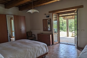 Ein Schlafzimmer mit einem Bett, einem Holzschrank, einem Schreibtisch und einem Stuhl. Eine Tür führt in einen Außenbereich mit Blick auf Bäume.