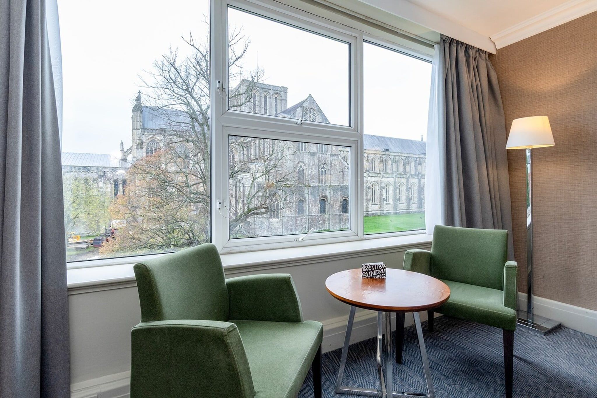 A hotel room with a large window, two green chairs, a small round table, and a floor lamp.