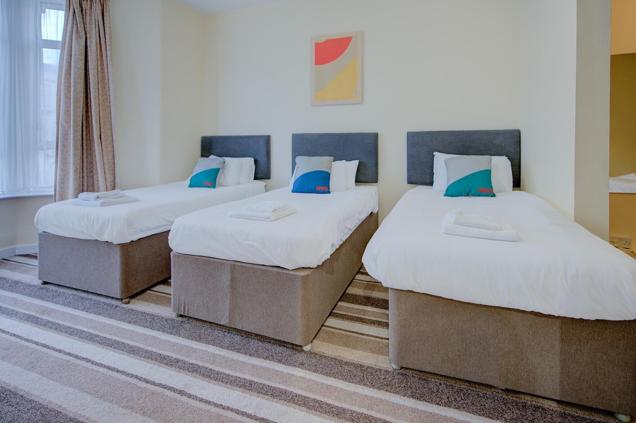 Une chambre d’hôtel avec trois lits, chacun doté d’une tête de lit grise et de draps blancs. Le sol est recouvert d’un tapis à rayures, et une fenêtre avec des rideaux se trouve sur le côté gauche.