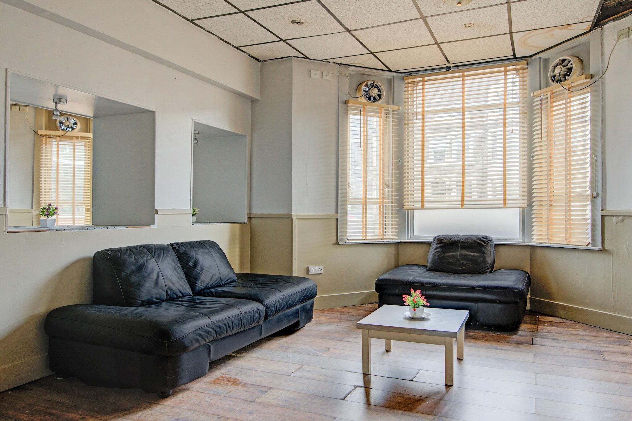 A living room with a black leather sofa, a smaller black sofa, and a white coffee table. There are two windows with blinds, a ceiling fan, and a mirror on the wall.