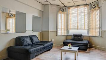 A living room with a black leather sofa, a smaller black sofa, and a white coffee table. There are two windows with blinds, a ceiling fan, and a mirror on the wall.