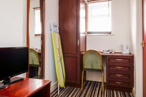 A hotel room with a wooden desk, a television, a mirror, a wardrobe, and a window with blinds.