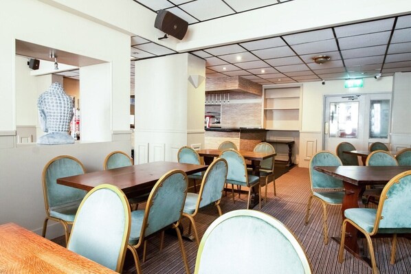 A dining area with multiple tables and chairs, a kitchen in the background, and a speaker on the ceiling.