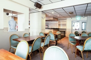 Un espace repas avec plusieurs tables et chaises, une cuisine à l’arrière-plan et un haut-parleur fixé au plafond.