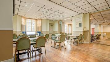 A spacious room with a high ceiling, multiple tables and chairs, and large windows with blinds.