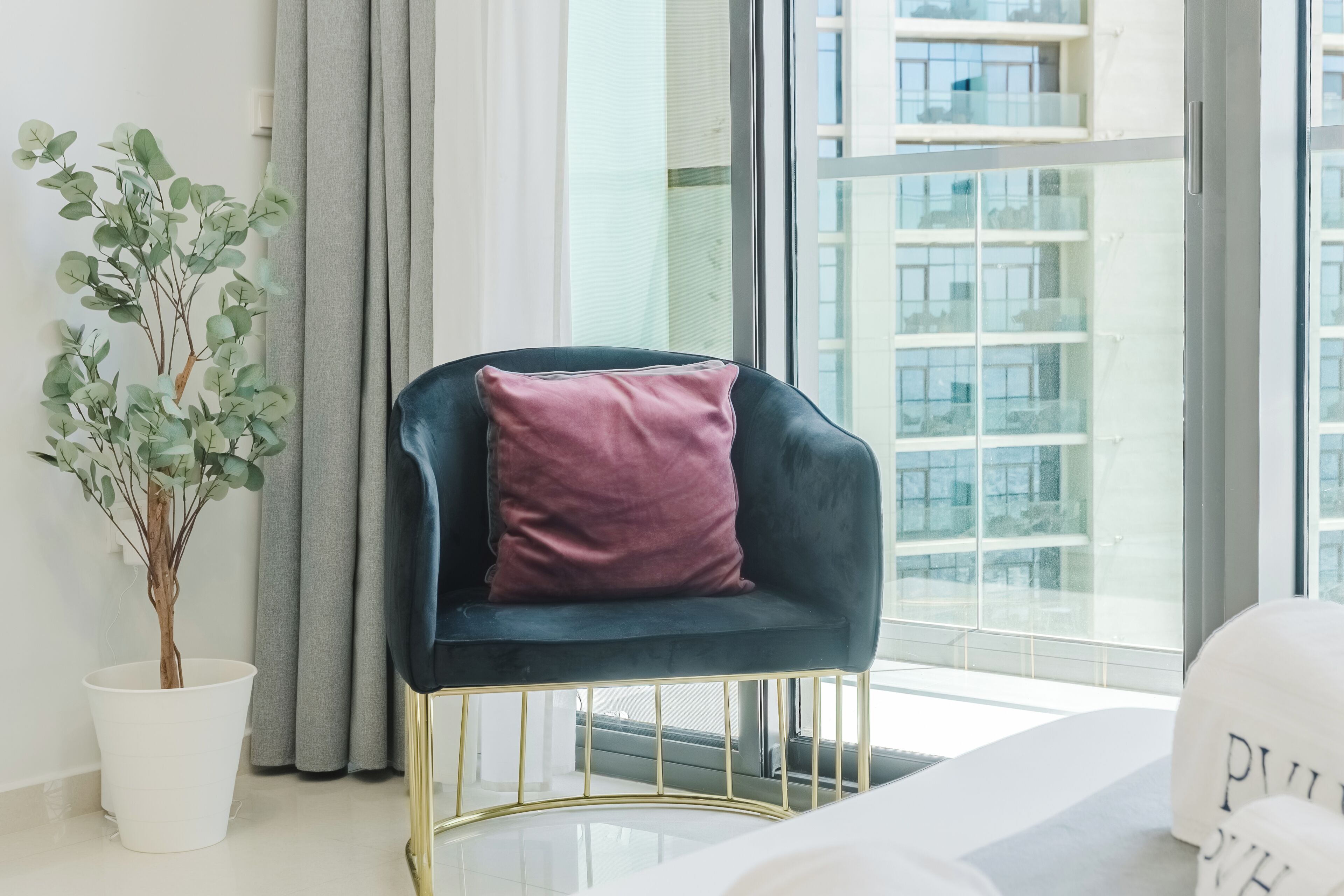 Una habitación moderna con una silla azul oscuro, una planta en maceta blanca y vistas a un edificio a través de amplias ventanas.