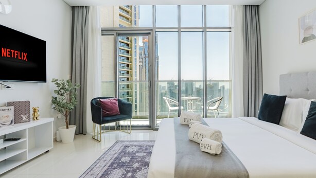 A modern hotel room with a large bed, a TV, a balcony with a city view, and a dining area. - PVH - Aykon City - Canal View Studio  (Dubai)