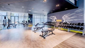 A modern gym with various exercise equipment, large windows, and a wall with a winged logo. - PVH - Aykon City - Canal View Studio  (Dubai)