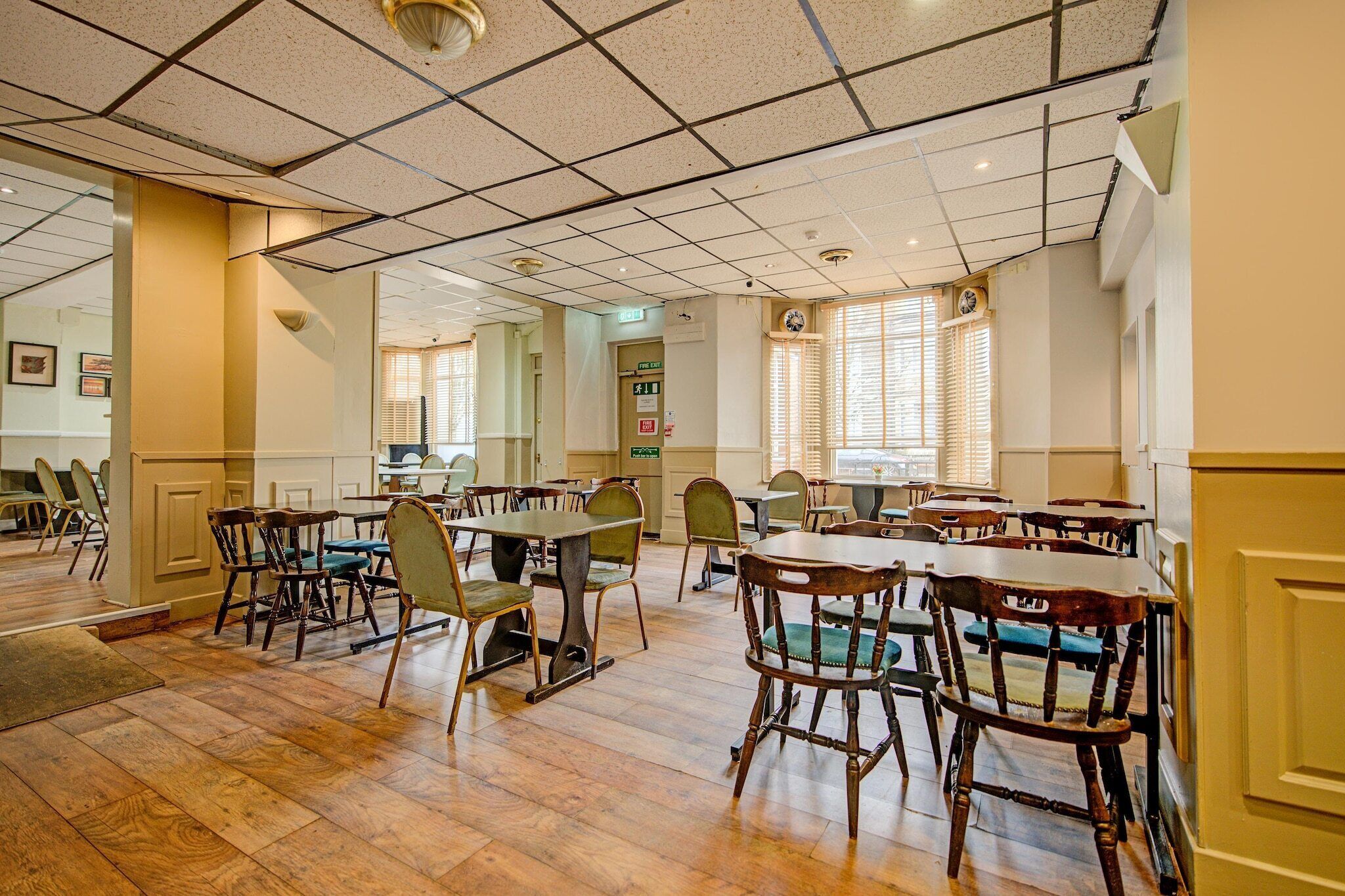 A dining area with wooden tables and chairs, a tiled ceiling, and a view of the outside through windows.