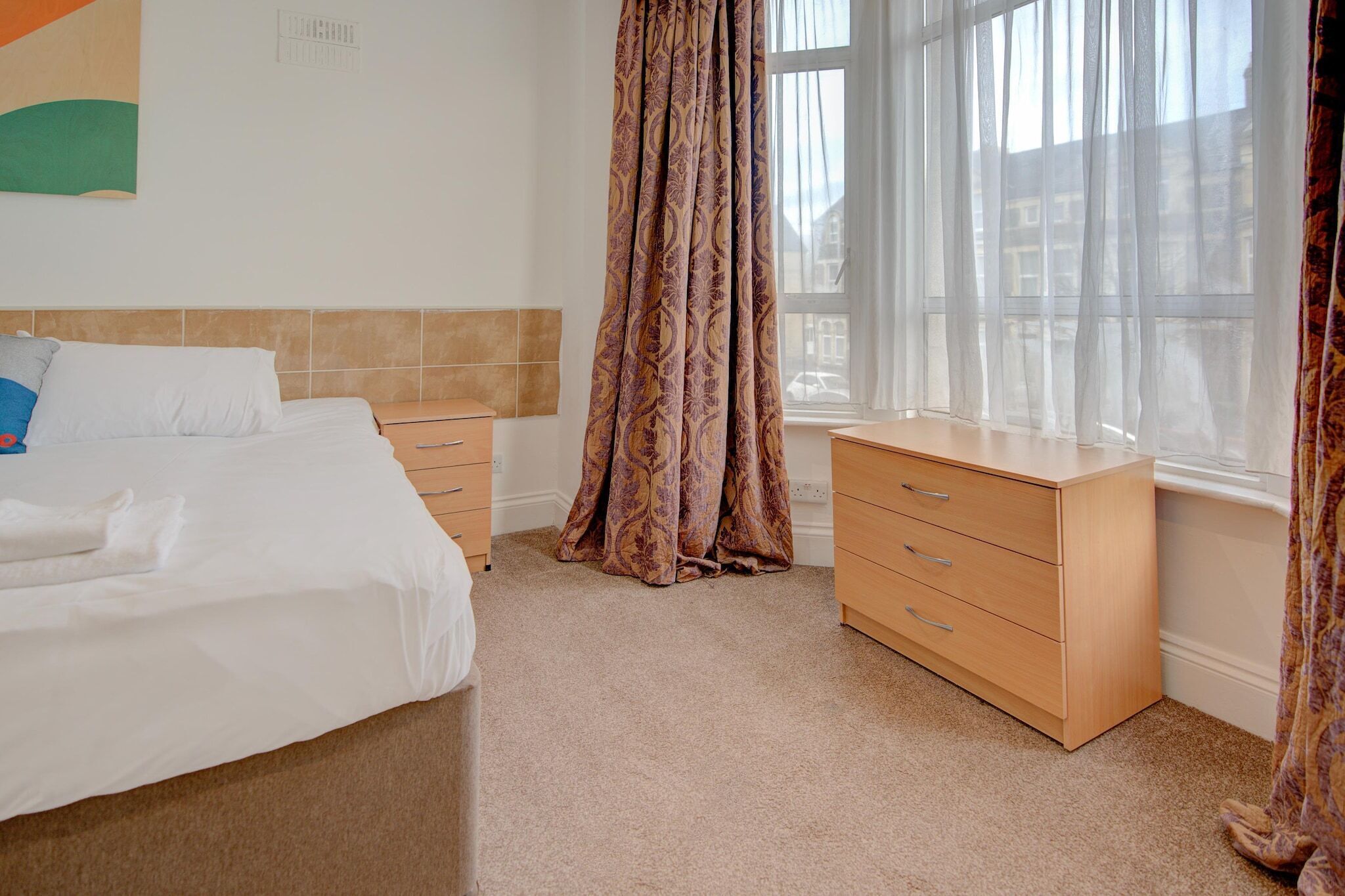 A hotel room with a bed, a wooden dresser, and a window with curtains.