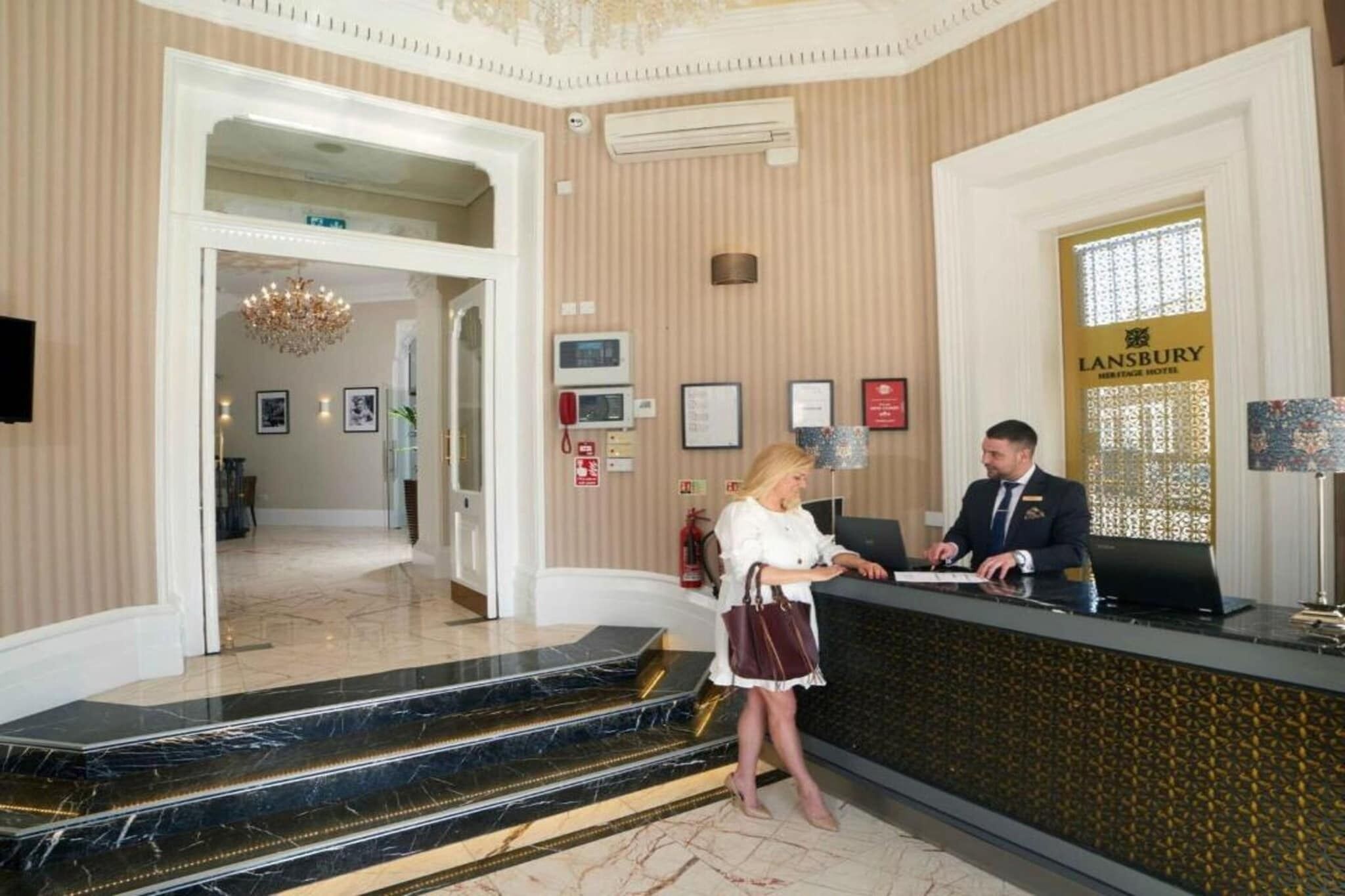 A hotel reception area with a reception desk, a chandelier, framed pictures, and a sign for Lansbury Heritage Hotel.