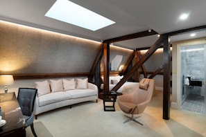 Een moderne woonkamer met een bank, fauteuil en een tafel. Er is een trap met houten balken en een dakvenster.