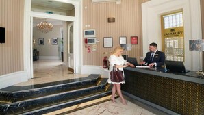 A hotel reception area with a reception desk, a chandelier, framed pictures, and a sign for Lansbury Heritage Hotel. - Lansbury Studio Suite (London)
