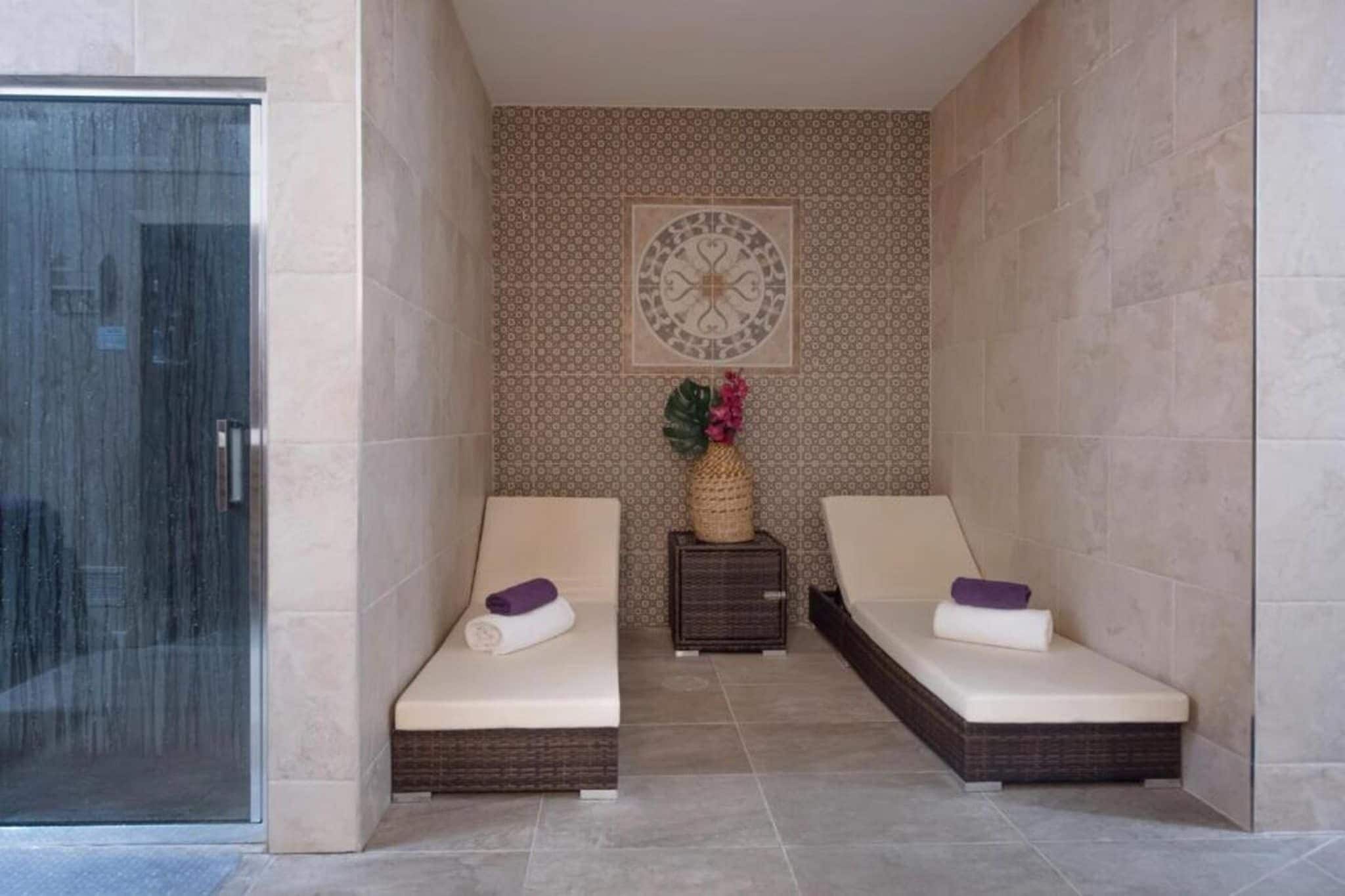 A spa room with two massage beds, a decorative wall, and a small side table with a plant.