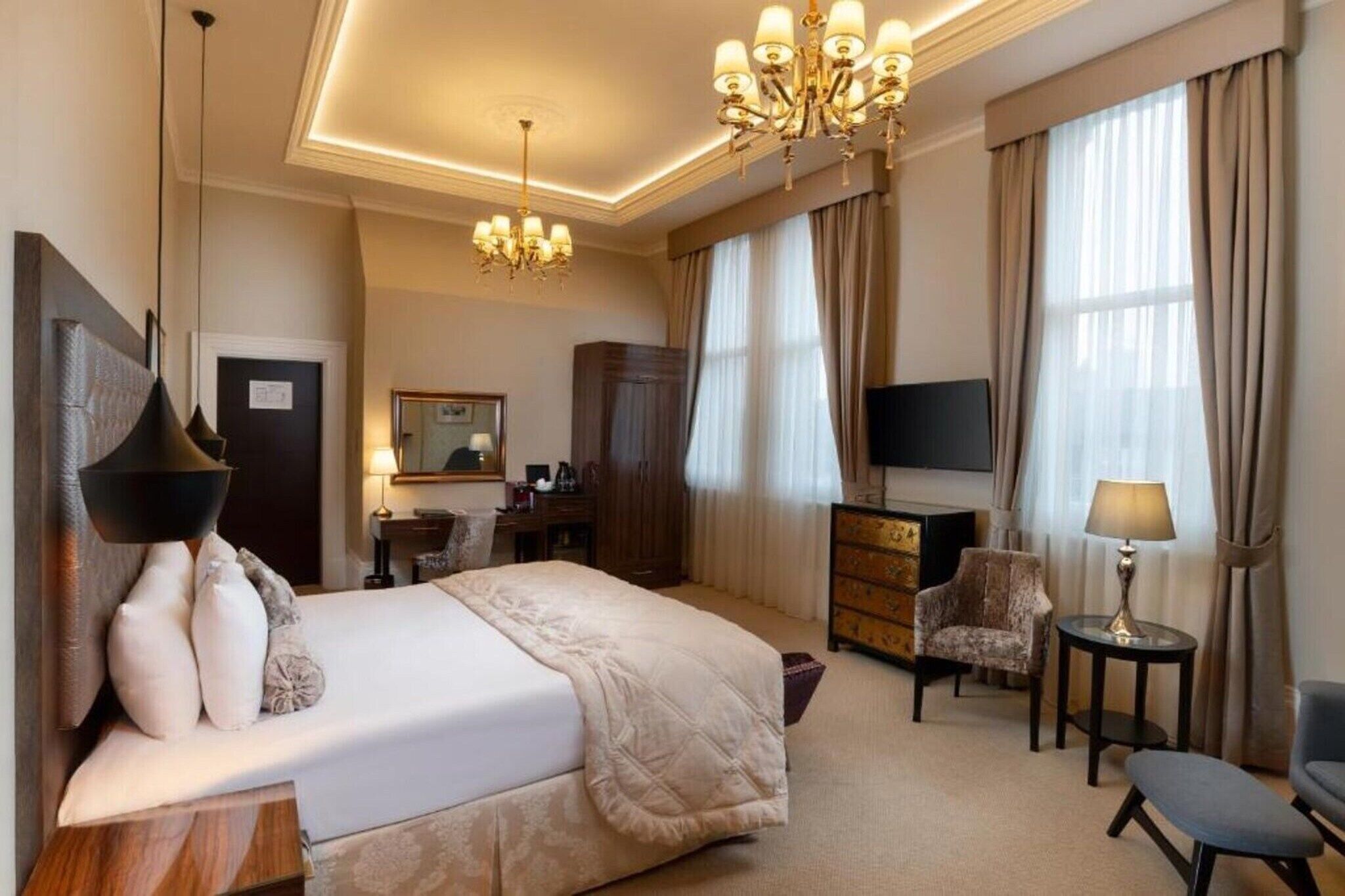 A hotel room with a large bed, a TV, a desk, a chair, and a chandelier.