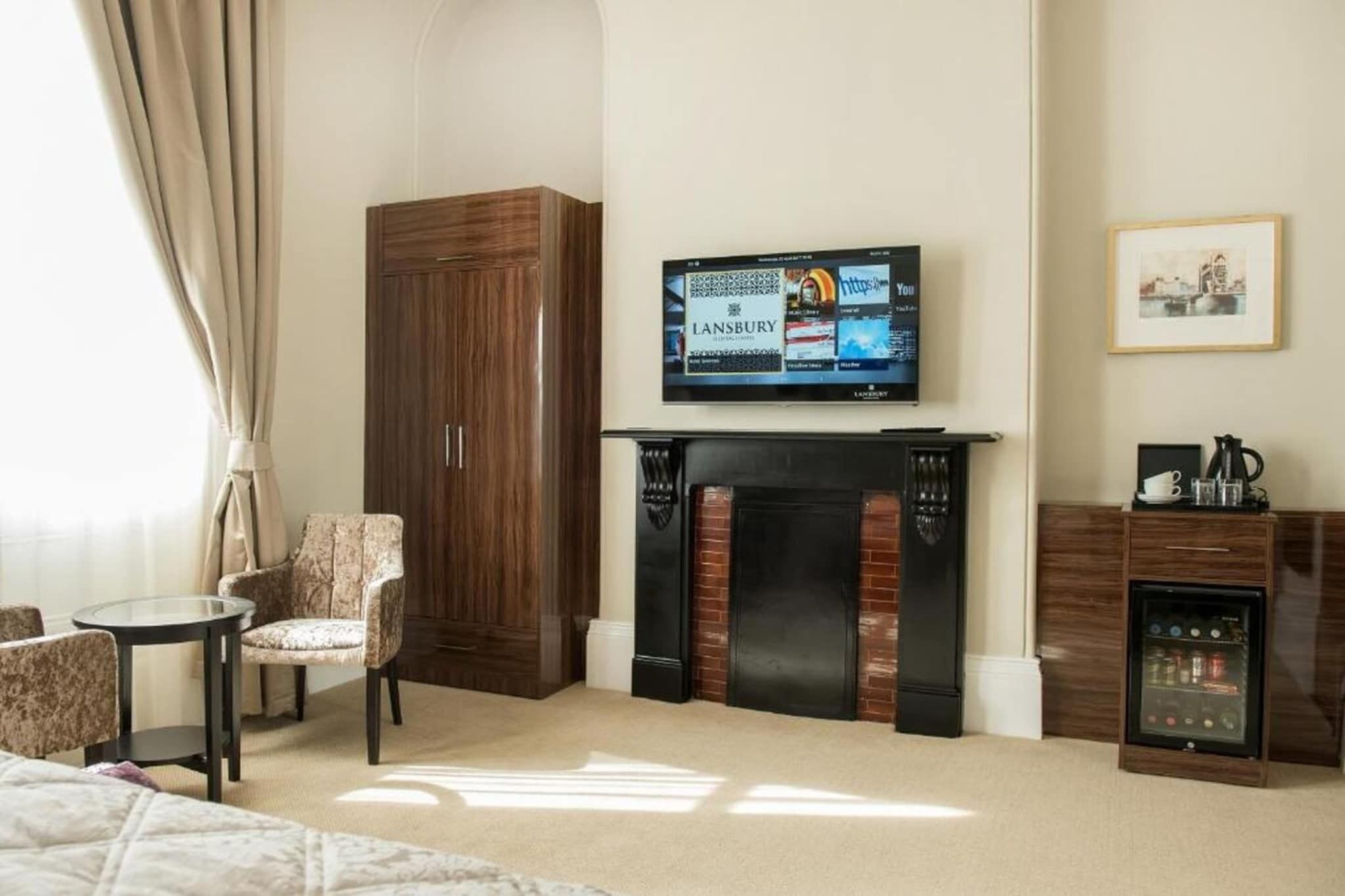 A hotel room with a television mounted above a fireplace, a seating area with a chair and a small table, and a bed with a headboard.