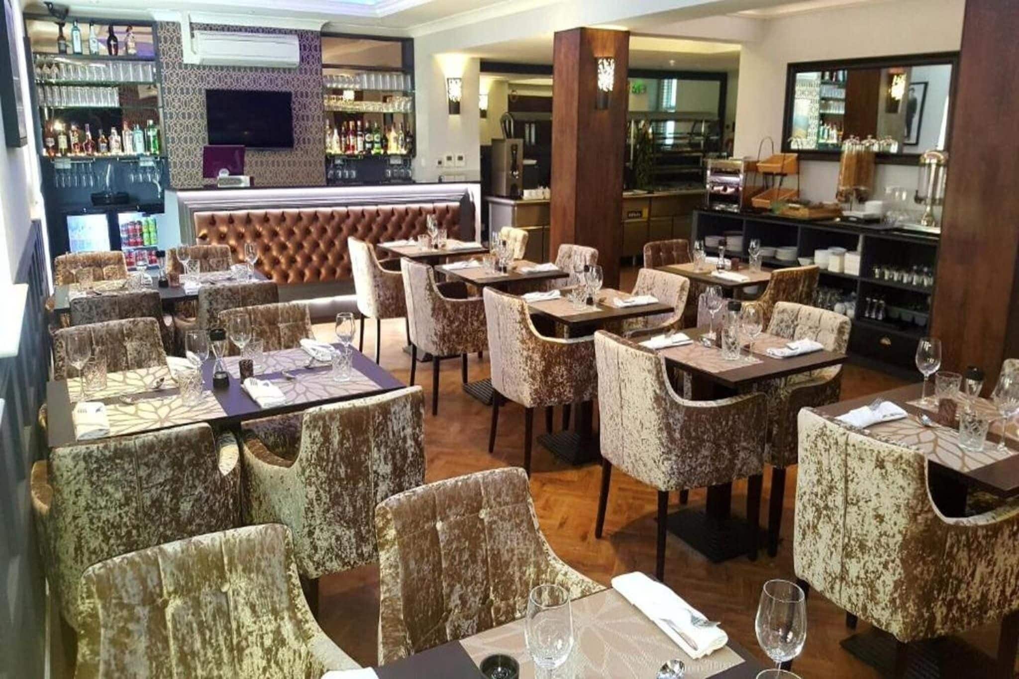 A restaurant with multiple dining tables and chairs, a bar area with shelves of bottles, and a television screen.