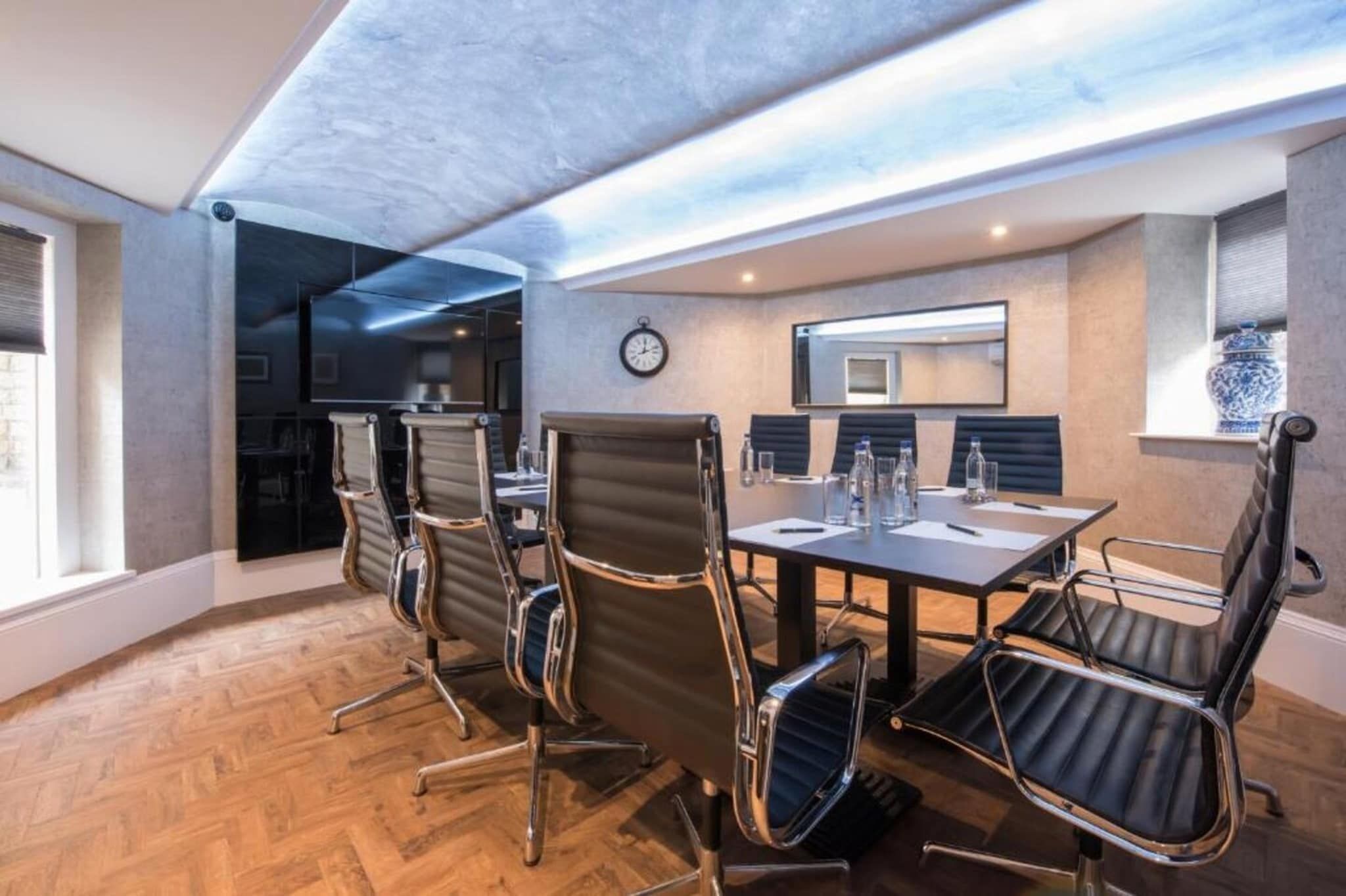 A conference room with a long table, six chairs, a clock, and a window with a vase.