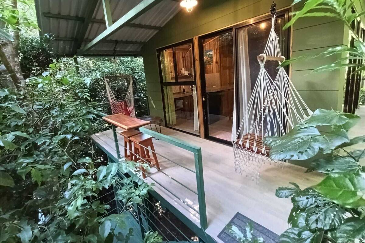 Un espacio exterior cubierto con una mesa y sillas de madera, una silla colgante de macramé y vistas del interior a través de una puerta de cristal.