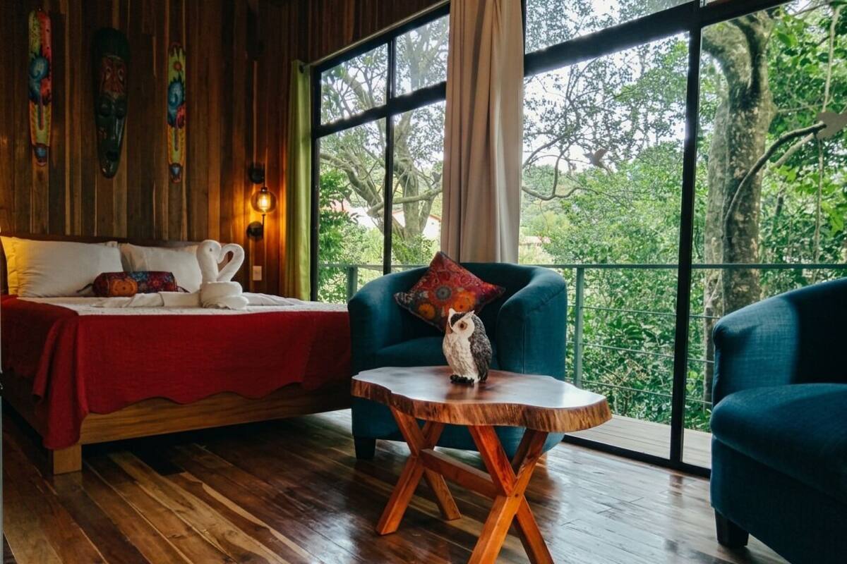 Una habitación con piso de madera, una cama, una mesa redonda de madera y dos sillones azules.
