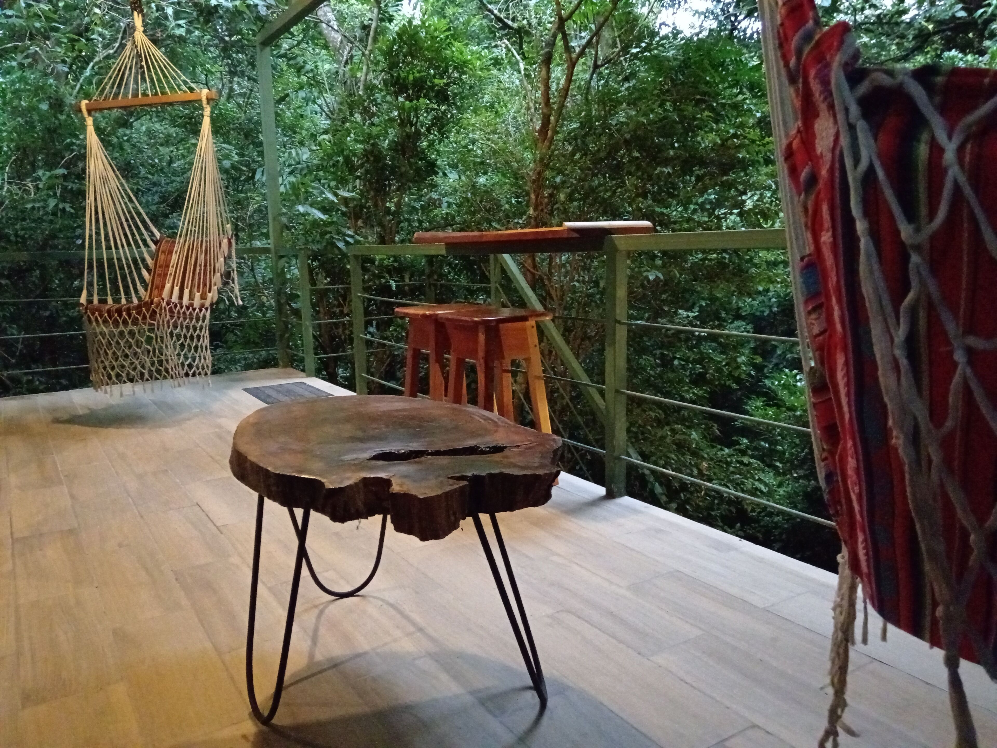 A hammock chair, a wooden table, and a red patterned fabric on a balcony with a view of dense greenery.