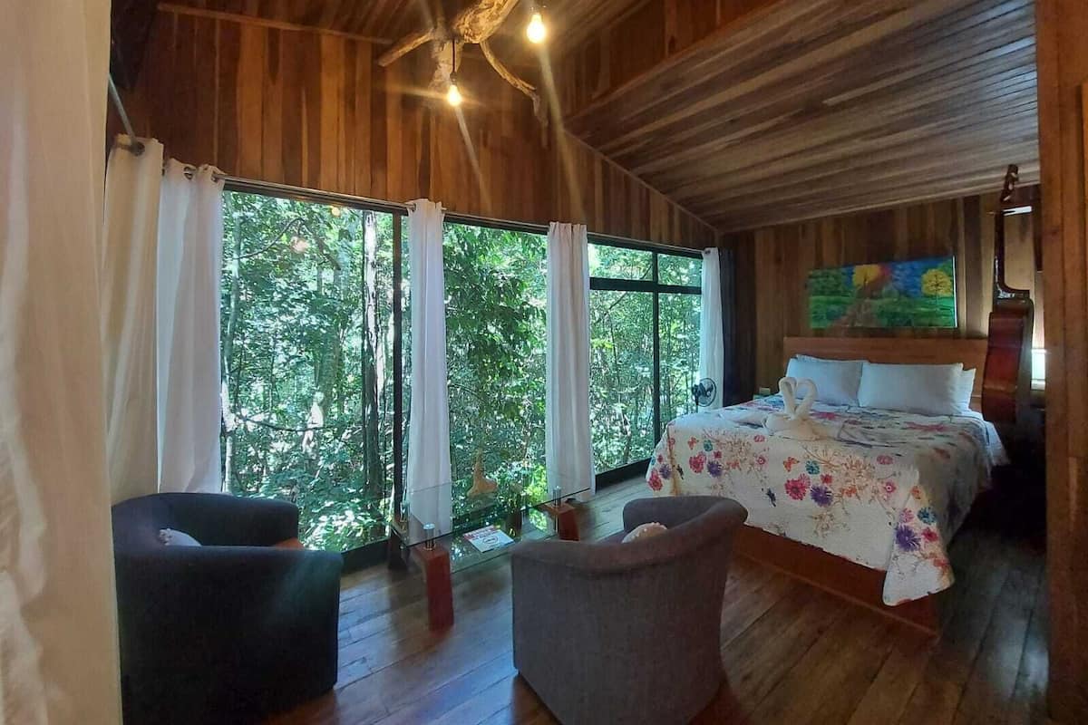 Una habitación de madera con una cama, dos sillas y ventanas grandes.