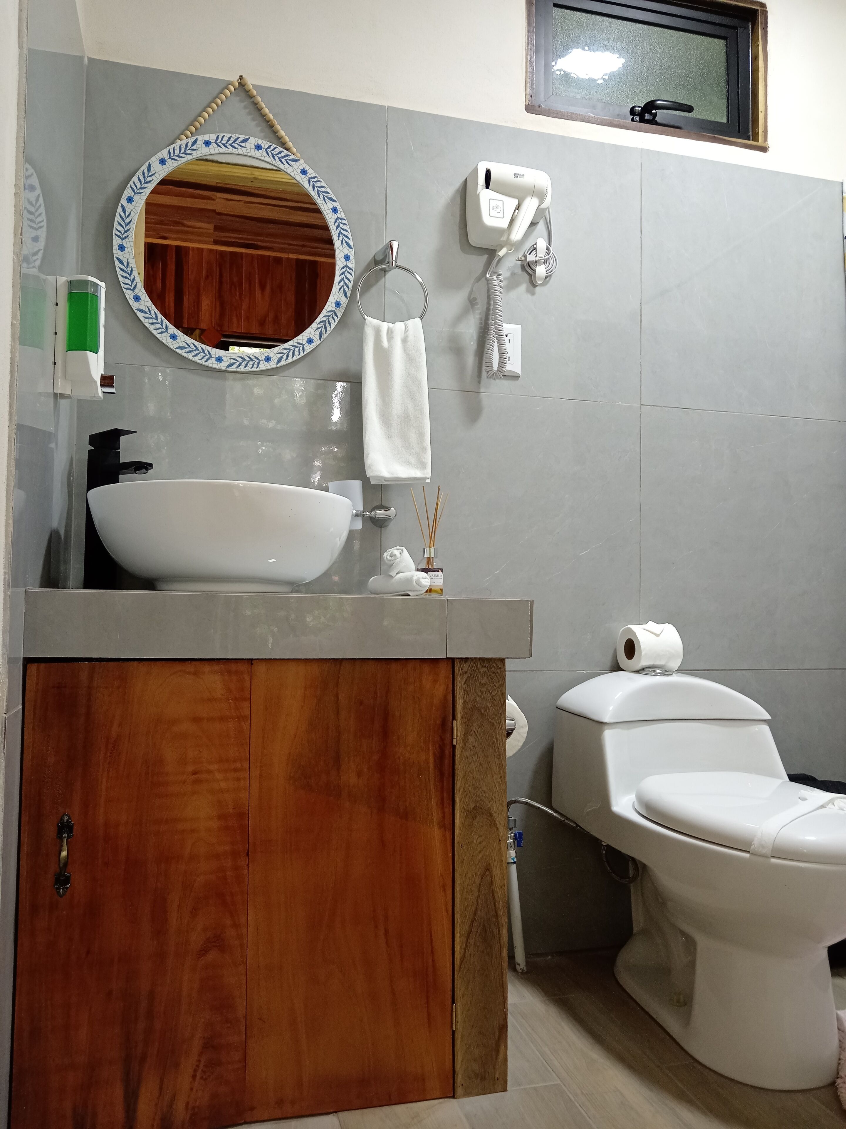 Une salle de bain avec un meuble-lavabo en bois, un lavabo blanc, des toilettes, un miroir avec un cadre décoratif, un porte-serviettes et un distributeur de savon.