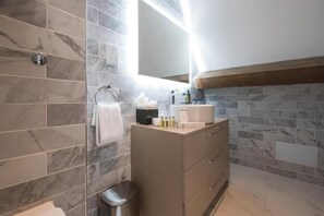 Un bany amb rajoles de marbre, un lavabo amb mirall, un suport per a tovalloles i un safata de brossa.