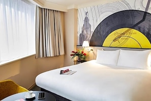 Una habitació d'hotel amb un llit gran, una taula de nit amb una làmpada, un ram de flors i una finestra amb cortines.