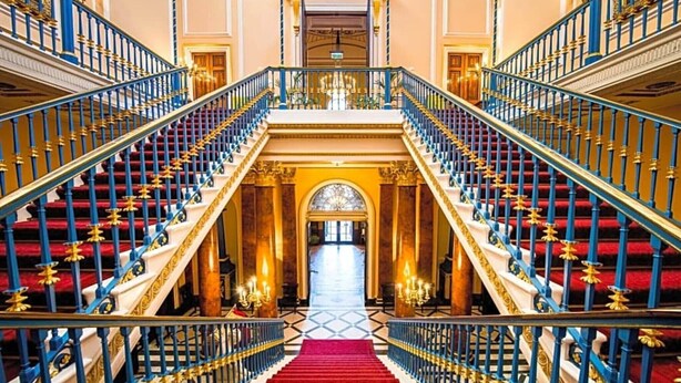 Een statige trap met rood tapijt en blauwe leuningen die leiden naar een centrale boog.