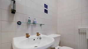 A bathroom with a white sink, two faucets, a toilet, and a glass shelf with toiletries.