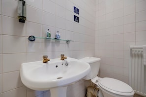 A bathroom with a white sink, two faucets, a toilet, and a glass shelf with toiletries.