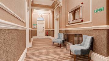 A hotel hallway with a carpeted floor, two armchairs, a fire exit sign, and a door labeled 'The Director's Suite.'