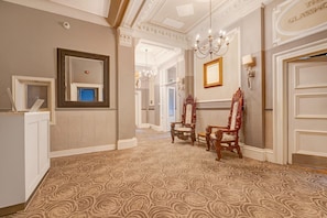 A hallway with ornate wall moldings, a chandelier, and two chairs with intricate designs.