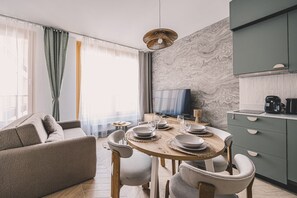Moderni olohuone, jossa on sohva, ruokapöytä, tuolit, televisio sekä keittiöalue, jossa on kaappeja ja kodinkoneita.