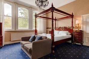 A four-poster canopy bed with white linens, a wicker sofa with patterned cushions, and a classic wooden nightstand with a lamp.