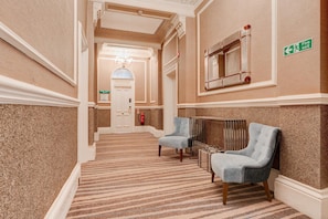 A hotel hallway with a carpeted floor, two armchairs, a fire exit sign, and a door labeled 'The Director's Suite.'