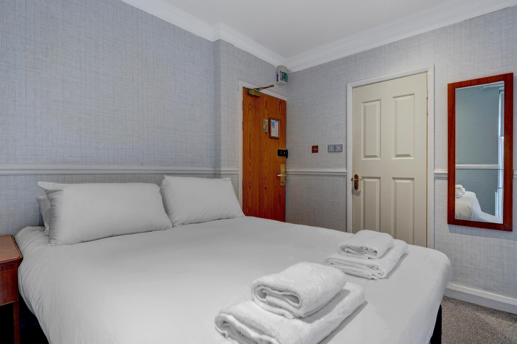 A hotel room with a large bed, a wooden door, a mirror, and a wall-mounted light fixture.