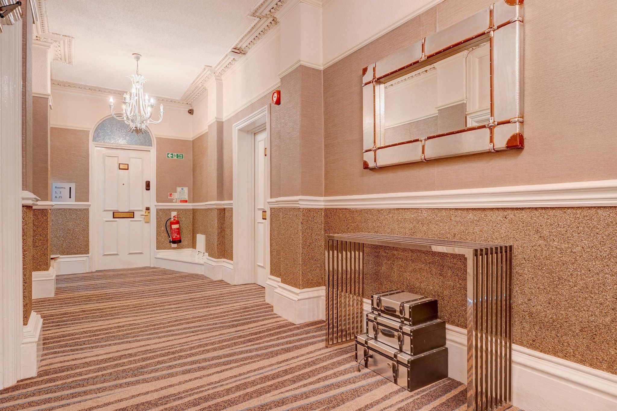Un hall d’hôtel avec un grand escalier, un lustre, un extincteur et un miroir au mur.