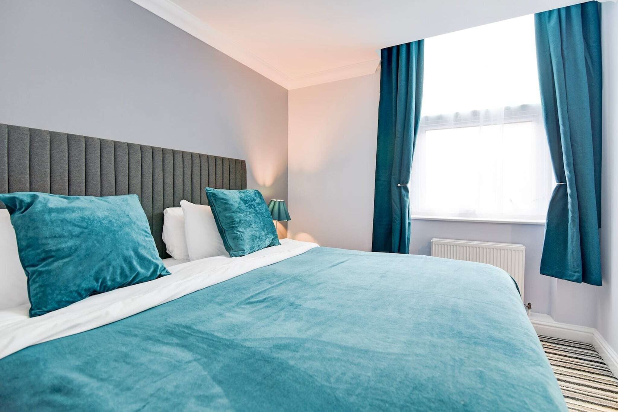 A modern hotel room with a large bed, teal bedding, a grey headboard, and a window with blue curtains.
