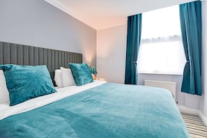 Une chambre d’hôtel moderne avec un grand lit, une literie de couleur turquoise, une tête de lit grise et une fenêtre agrémentée de rideaux bleus.