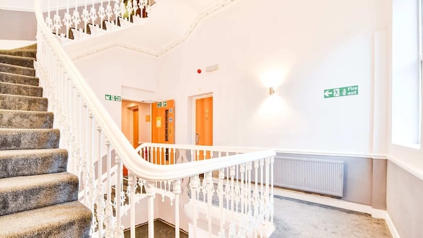 Un escalier avec une rampe blanche, des marches recouvertes de moquette et un mur portant un panneau indiquant la sortie de secours.